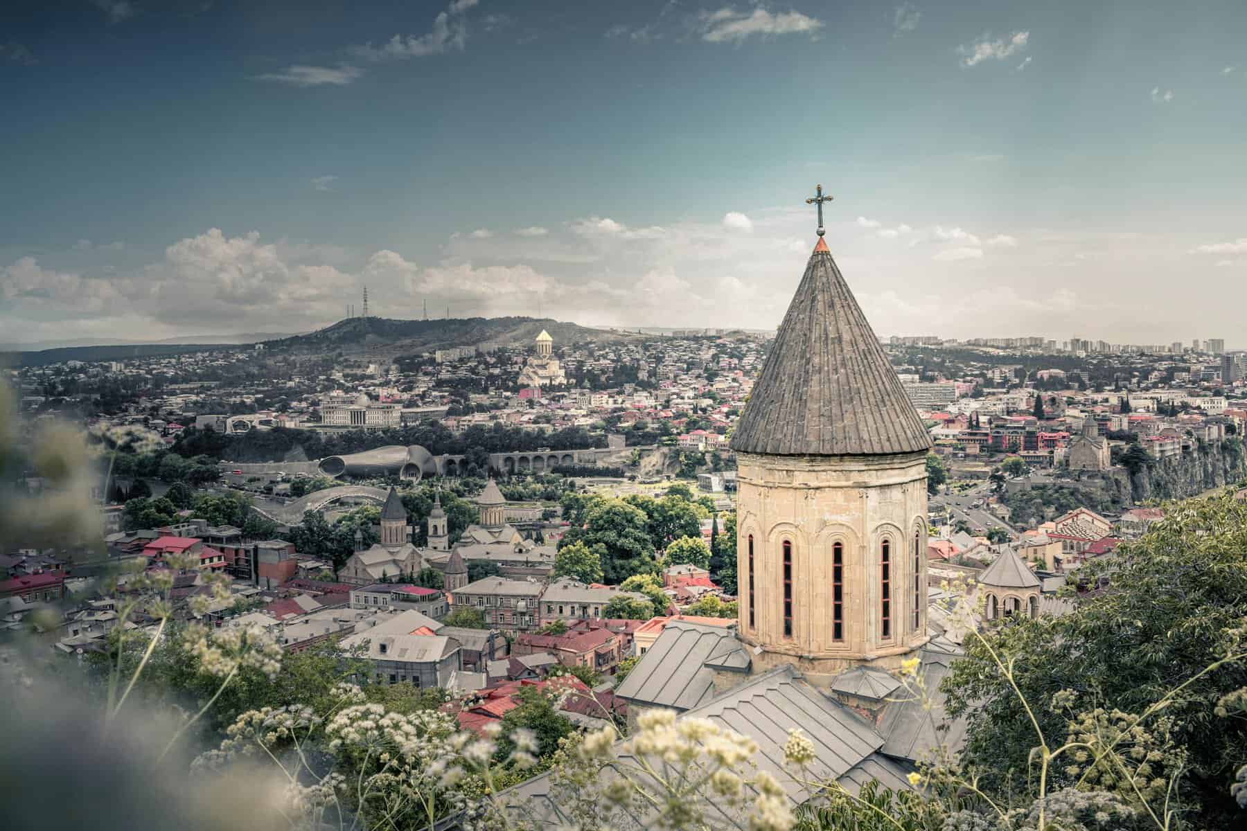 View of a historical church tower with Tbilisi's cityscape in the background under a blue sky.